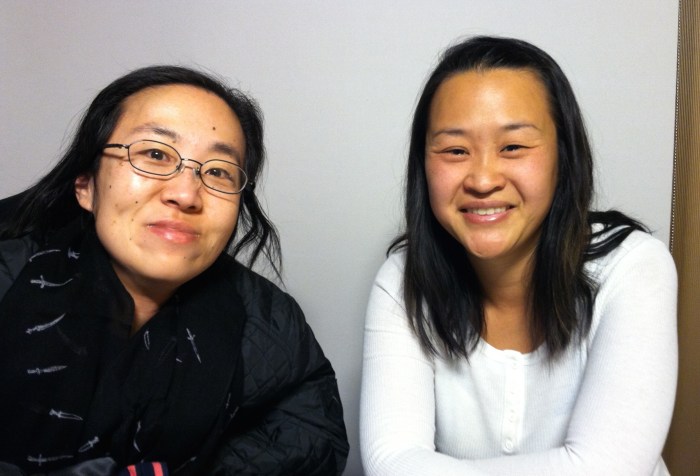 Two Asian American women sitting side-by-side smiling at the camera. The woman on the right is wearing a black jacket and glasses. The woman on the right is wearing a white shirt and has long black hair.