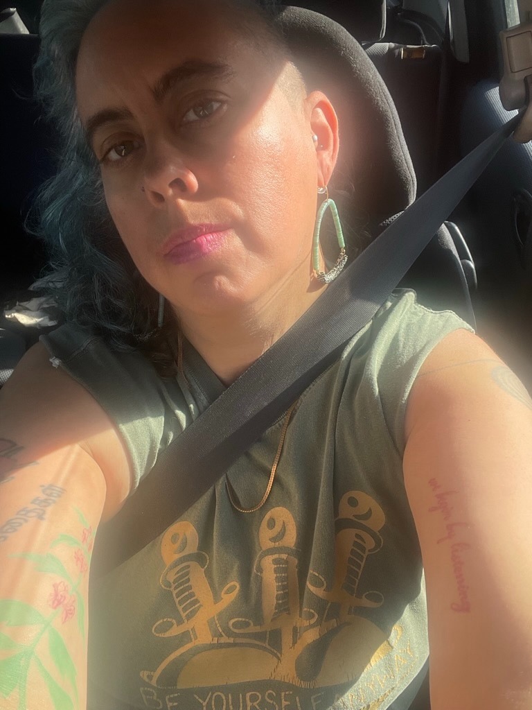 a nonbinary Sri Lankan/ white femme smirks at the camera, they are wearing an olive green cut off shirt with three swords poking into a heart and a gold chain necklace, blue and brown curly hair with a fresh silver side shave, looking speculatively at the viewer while seated in a car