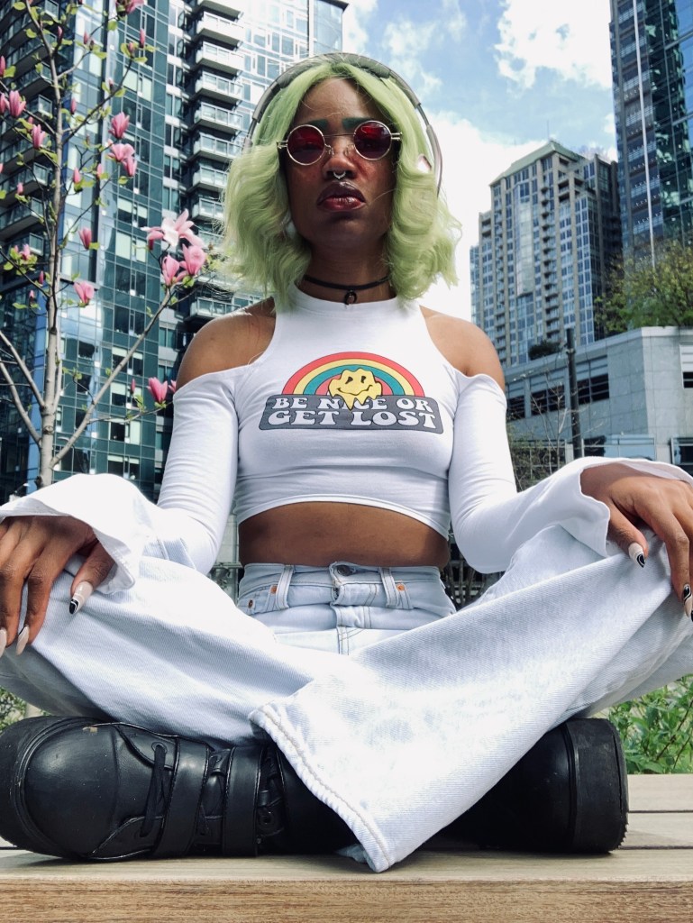 Image of Faati, a nonbinary light brown skinned Black Native, sitting cross legged in front of a cherry blossom tree and multiple high rise buildings. They have on acid washed light blue bell-bottom jeans, a white crop topped long sleeved shirt and black platform boots. They are sitting cross-legged with their hands on their knees. Their fingernails are a long almond acrylic style with nude coloring and black and white designs on top. They have green straight 70s styled hair with the ends bumped. They are wearing circle 70s style red lens sunglasses.