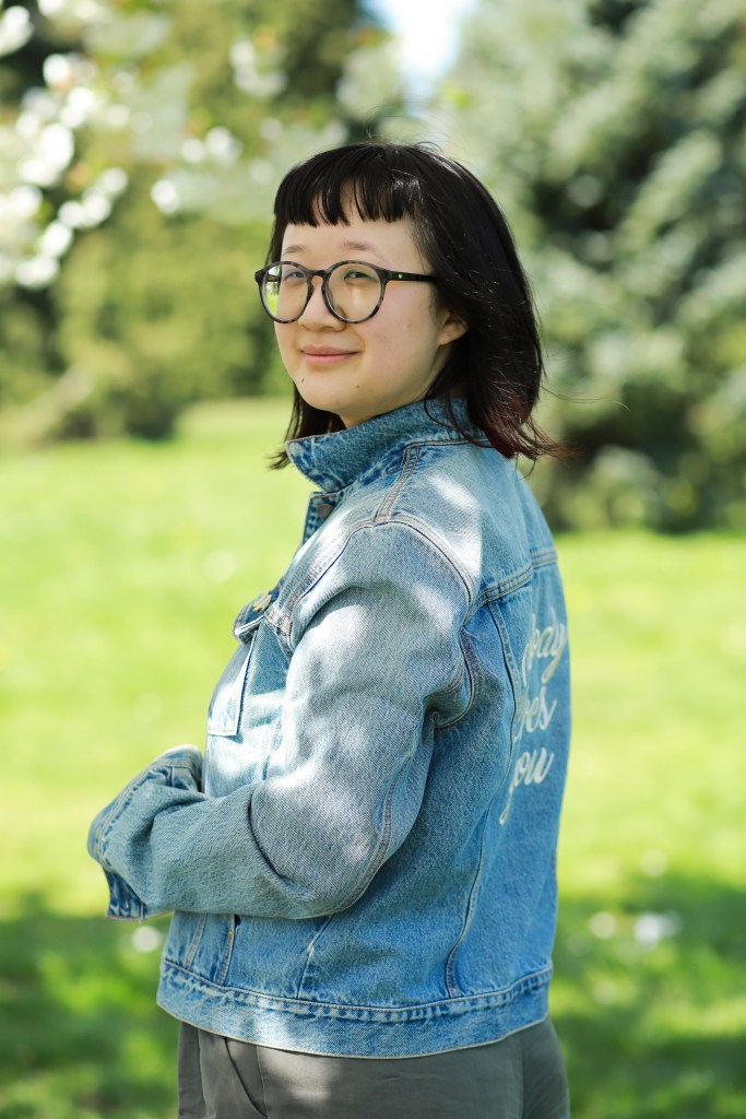 A person with pale-tan skin with her body away from the camera but her face smiling towards it. She's wearing a blue jean jacket that reads "Nobody Loves You" in the back, embroidered in white calligraphy. Her hair is nearly shoulder-length with dark red highlights at the end and she's wearing large round black-gray glasses. The background is a park during the day-time. Photo credit: Joy Gyamfi