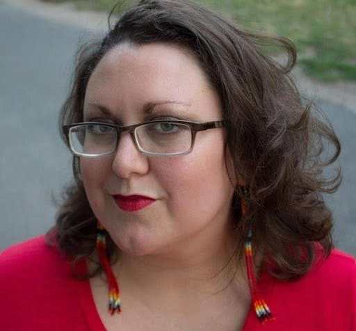 Close up of a light complexion, Two Spirit femme with black glasses, green eyes, shoulder length brown hair, Native style beaded dangling earrings, with bright red lips and a red shirt. In the foreground is a paved path and grass.
