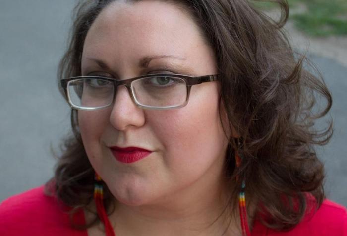 Close up of a light complexion, Two Spirit femme with black glasses, green eyes, shoulder length brown hair, Native style beaded dangling earrings, with bright red lips and a red shirt. In the foreground is a paved path and grass.