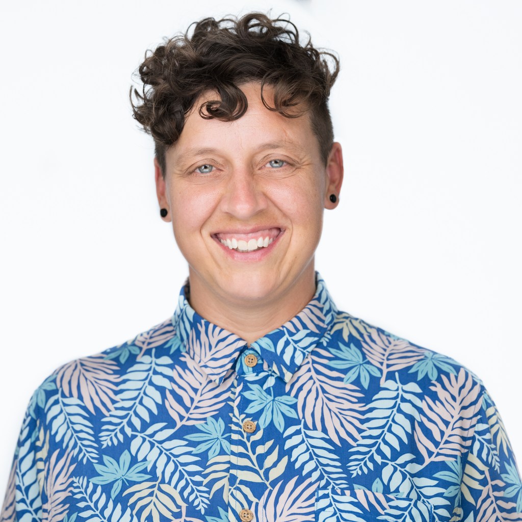 A smiling non-binary white person with short curly brown hair wears black earring plugs and a bright blue button up shirt with colorful fern fronds printed on it.