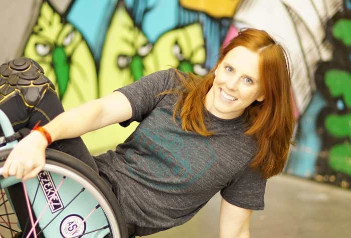 Katherine Beattie, a young white woman with long red hair. She is wearing a gray t-shirt and in her wheelchair popped at a 45 degree angle against a colorful mural.