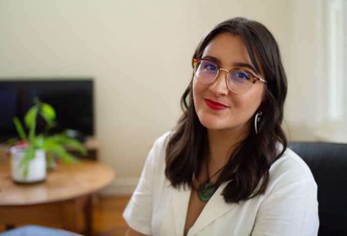 Photo of Kera Sherwood-O'Regan, an Indigenous Māori woman with light olive skin and long brown hair, sitting in her living room. She is wearing a white collared jumpsuit, vintage style tortoiseshell glasses, Haus of Dizzy love heart earrings, and red lipstick. She wears a Māori pounamu greenstone pendant around her neck, and she is smiling slightly at the camera. There is a green houseplant and wooden furniture in the background.