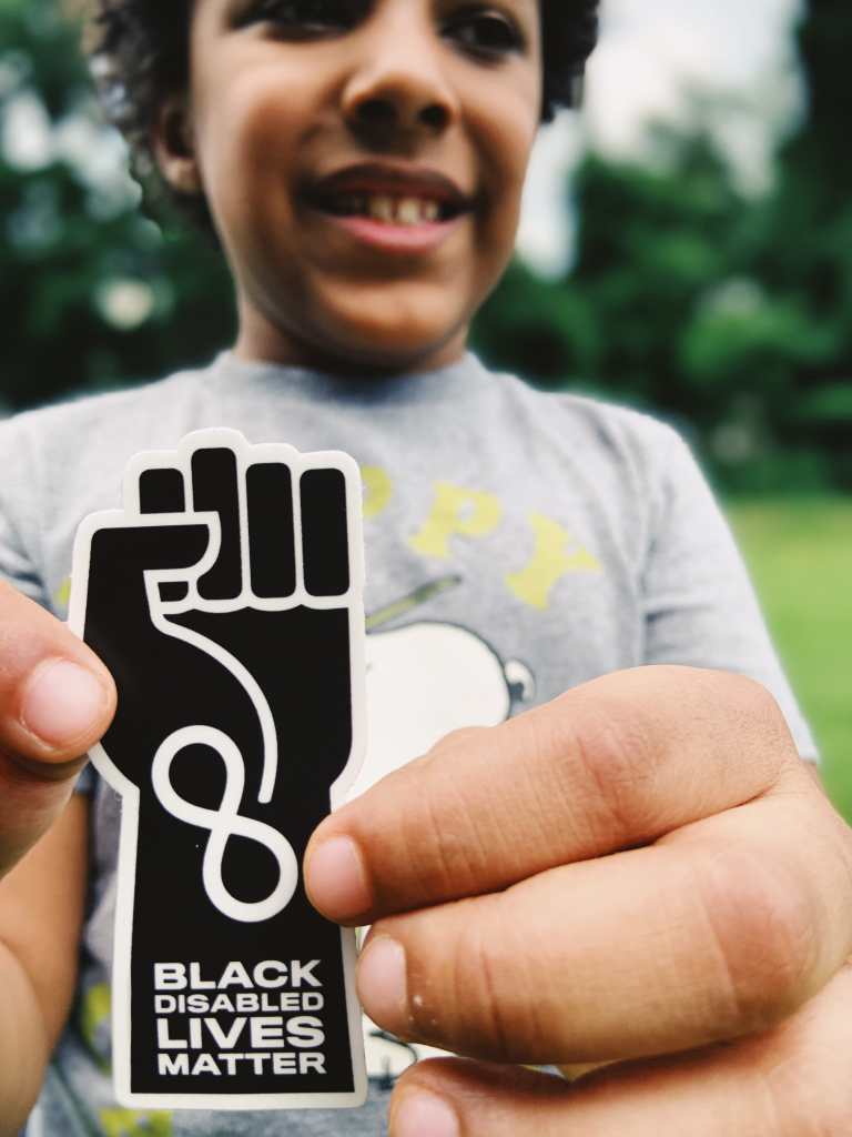 Photo of Knox White-Johnson, a Black Autistic boy smiling and holding a sticker designed by his mother, Jen White-Johnson, an illustration of a raised solidarity fist in black with an infinity symbol at the wrist in white with the words ‘Black Disabled Lives Matter’ below.