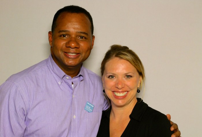 African American man with short hair wearing a light purple shirt. Next to him is a white woman with blonde hair wearing a black shirt. They are both smiling at the camera.