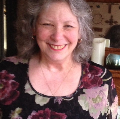 An older white woman with long gray hair. She is wearing a black top with flowers.