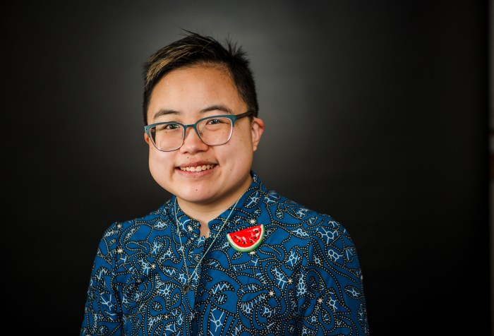 Ly Xīnzhèn is an androgynous and transmasculine East Asian person in their early thirties. They have short black hair with light highlights and they’re wearing a blue top with an abstract pattern, a handmade watermelon pin, and a silver magen david necklace.