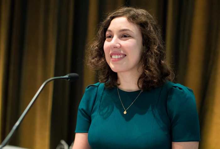 A white woman with curly brown hair is standing at a microphone, smiling. She is wearing a green dress and a simple gold necklace. Photo credit: John Livzey.