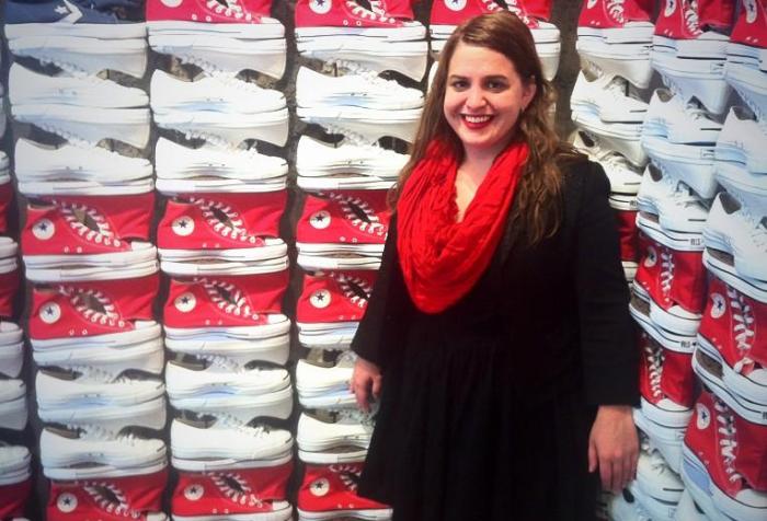 Young white woman with long brown hair who is wearing a black dress. She is standing behind a display of rows of sneakers that are red, white and blue.