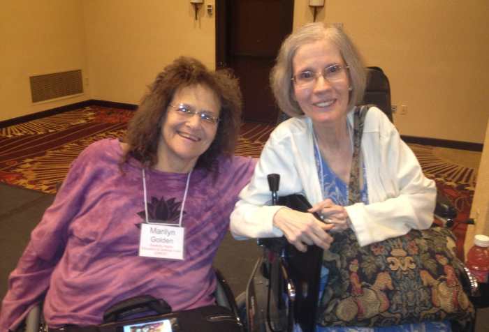 Marilyn Golden [left] in her manual wheelchair with brown curly hair, wire rimmed glasses, purple shirt, conference name tag, a briefcase in her lap, seated next to Diane Coleman [right] in her motorized wheelchair with gray and blond bobbed hair, wire rimmed glasses, blue patterned dress with white sweater, tapestry shoulder bag and slim briefcase to her side. Both are smiling and leaning toward each other. The year is 2014.