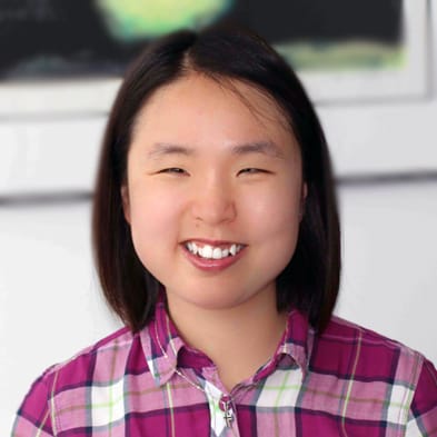 A photo headshot of a young woman with black hair smiling at the camera, wearing a white and plum colored plaid flannel shirt