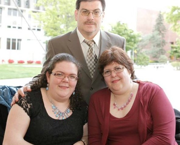Photo of three family members in a photo. In the front is a white young woman sitting in a wheelchair on the left. On the right, is a middle aged white woman in a wheelchair on the right. Behind the two women in a middle aged white man. All three have brown hair and are wearing glasses.