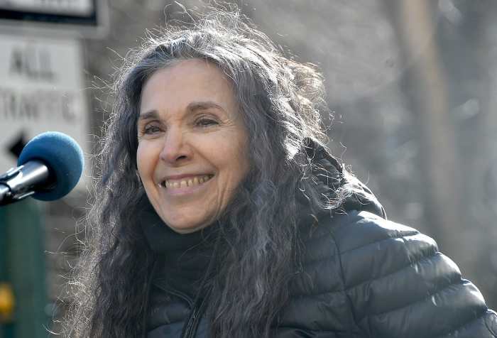 Photo of Nadia LaSpina, an Italian American woman with long curly gray hair with a microphone in front of her. She is wearing a black puffy jacket outdoors.