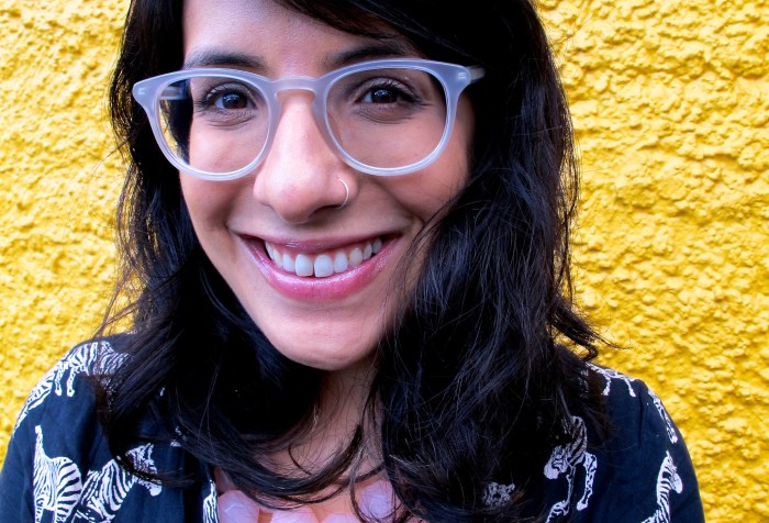Photo of Neera Jain, a smiling Indian-American woman with wavy shoulder-length brown hair and bangs, clear plastic-framed glasses, and a silver nose ring standing in front of a bright yellow stucco wall. She wears a black shirt-dress printed with white zebras, and a chunky pale pink beaded necklace.