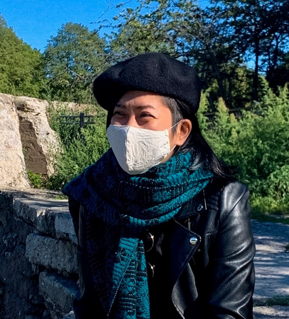 A photo of Akemi, East Asian woman with side shaved medium length black straight hair. She is wearing a black beret, green scarf, and a white mask. She is sitting outside in front of river and trees