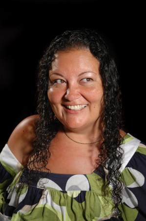 Image description: A woman wearing a green, white and blue blouse is centered in a photo. She is brown skinned with curly hair and is looking to the side with a large smile.