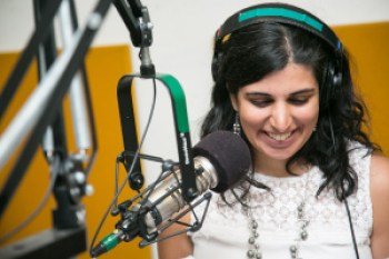 Photo of a South Asian American woman in front of a microphone at a radio station. She is wearing headphones and has long black hair. She is smiling and her eyes are downcast looking at something. She is wearing a white sleeveless shirt and a silver necklace. Photo credit: Ann Oleinik