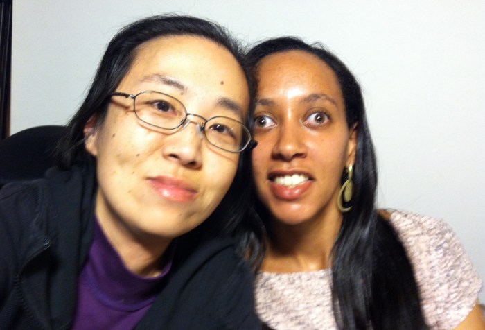 Asian American woman on the left in a black hoodie and wearing glasses. On the right is a young African American woman with long black hair and a short sleeve floral print top