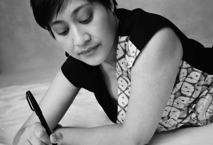 Black and white photo of an Indonesian woman with short hair, earrings, and a patterned dress, lying down on her front, pen in hand, ready to write. Pic credit: Derrick Kakembo.