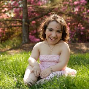 Cuquis sitting on the grass with her hands in her lap, smiling while wearing a light pink strapless dress, with a small rose gold heart necklace and low tan heels.