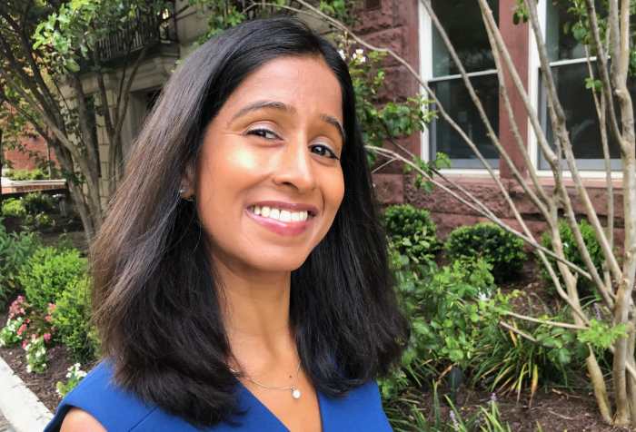 Qudsiya is wearing a blue dress and standing beside plants in front of a building on a city street.