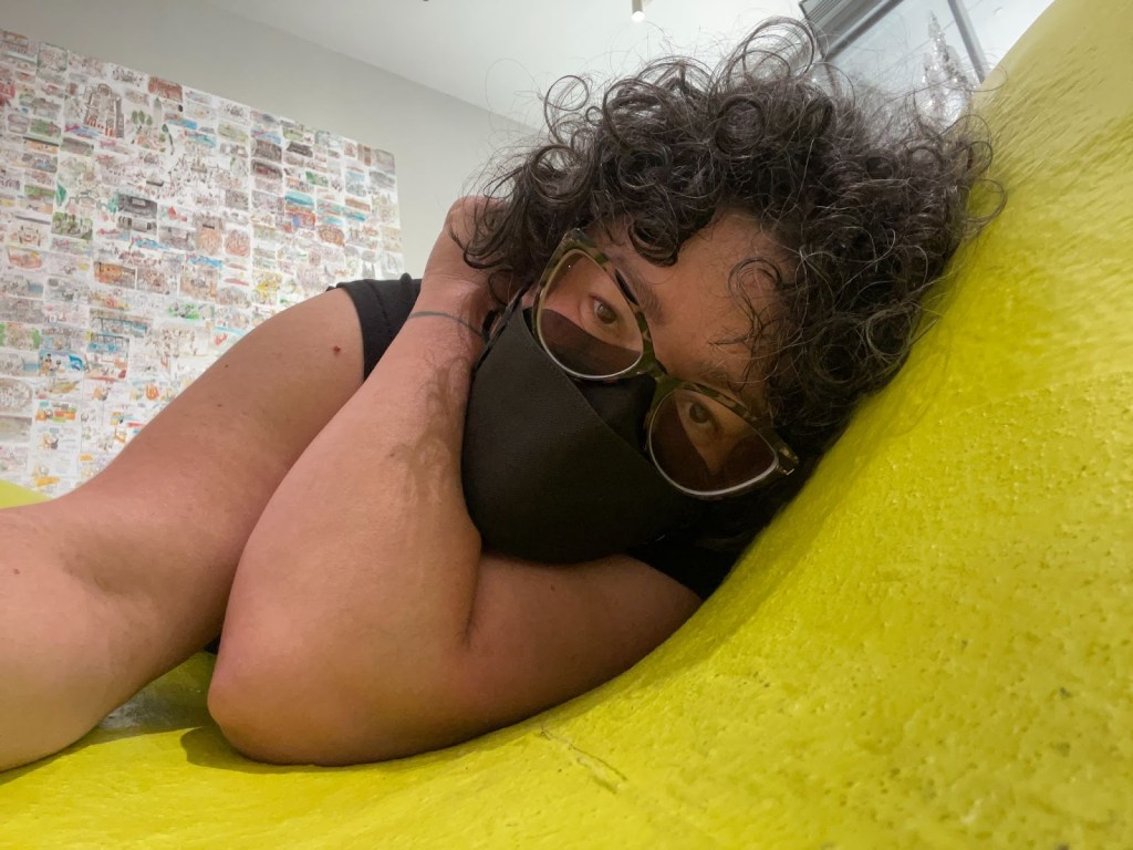 A selfie of Jillian Crochet, a white woman with tan skin and curly brown hair, wearing a black mask and tortoise shell glasses is laying against her chartreuse green Lumpy Bed sculpture/lounge. In the background is a wall of tiny sketches by the artist José Figueroa.
