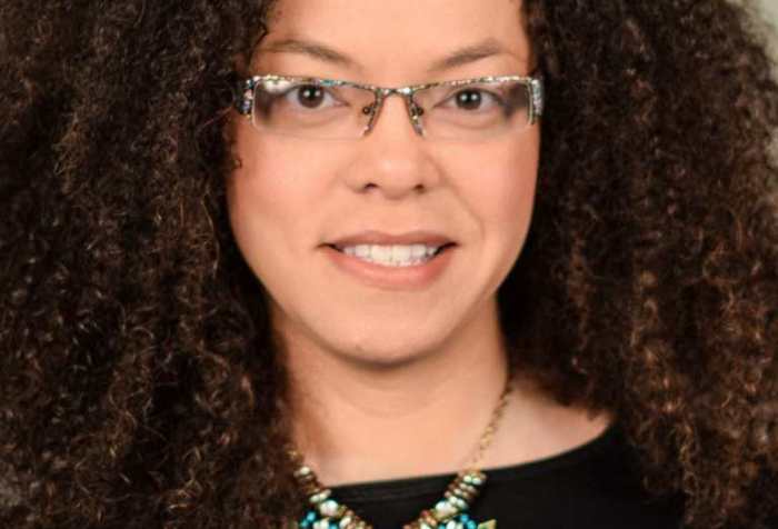 Headshot of a light complected African American woman, late thirties, with curly brown hair and glasses. She is wearing a black top and a large statement necklace