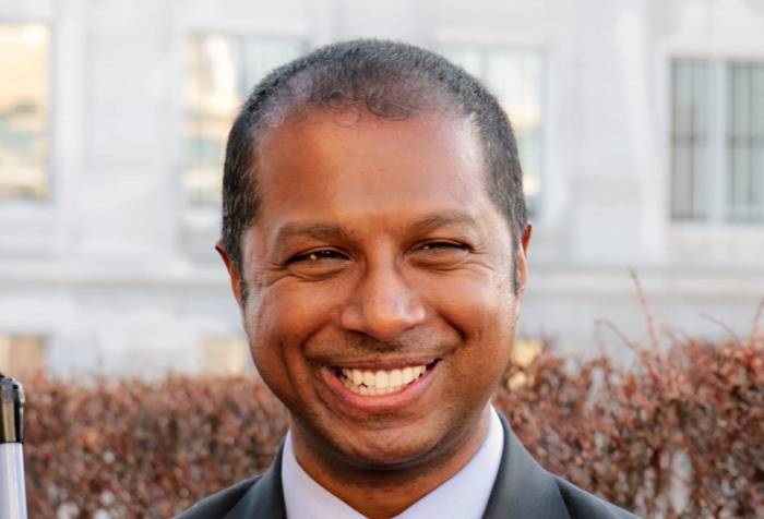 Picture of Sachin Pavithran, a South Asian man in a suit holding white cane outside the Capitol in Salt Lake City.