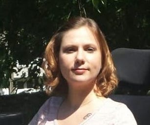 A young, white woman with short blonde hair is pictured outside. She is wearing a pink tee-shirt, black skirt with pink and green design, and matching heels. She is smiling toward the camera and seated in a black power wheelchair.