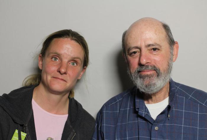 Image of two people standing next to each other. On the left is a young white woman with blonde hair tied back in a ponytail. She is wearing a black hoodie and pink t-shirt. To her right is an older white man with a gray beard. He is wearing a blue long-sleeved shirt.