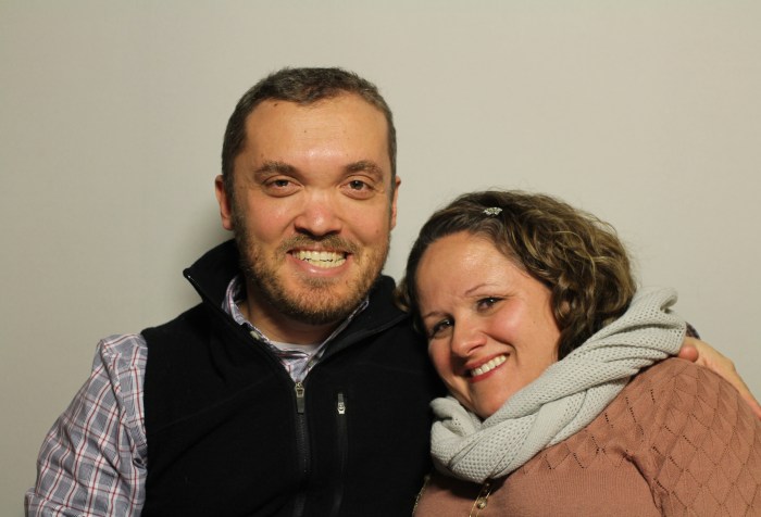 Image description: A photo featuring Eli Gelardin and Christina Mills taken on January 3, 2015. A close-up portrait of a person on the left who appears to be a white man with short brown hair and a beard. He is smiling and wearing a black vest and long-sleeved shirt. To his right is a person who appears to be a white woman with long wavy brown hair that has a barrette on the left side of her hair. She is smiling at the camera and bundled with a white knit scarf around her neck and a rose-colored sweater.