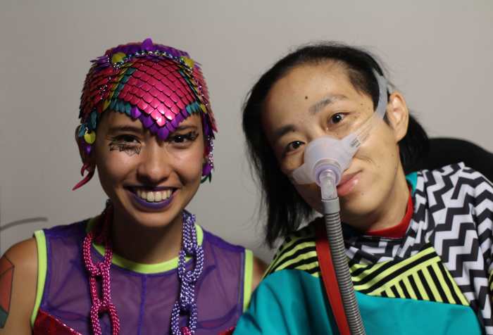 On the left is Sky Cubacub, a non-binary queer and disabled Filipinx human wearing colorful chainmaille headpiece in magenta, purple, turquoise and yellow. She is wearing a sleeveless purple mesh tank and a necklace that looks like chains in purple and magenta. On the right is Alice Wong, an Asian American disabled woman in a wheelchair. She is wearing a top with a geometric print with stripes, in aqua, black, white, and red. She is wearing a mask over her nose attached to a tube for her ventilator.