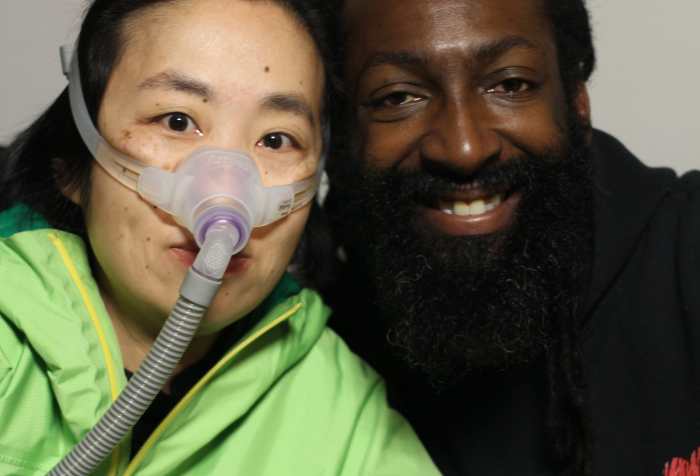 On the left, Alice Wong an Asian American woman wearing a bright green rain jacket. She is wearing a mask over her nose attached to a gray tube which is connected to her ventilator. She is sitting closely next to Antoine Hunter, a self-described handsome, dark milk chocolate, African American Man. He has long Ebony dreadlocks hair tied to back with a full Ebony-colored beard and full brown lips. He is wearing a black hoodie that says, “I [heart] Being BLACK.”