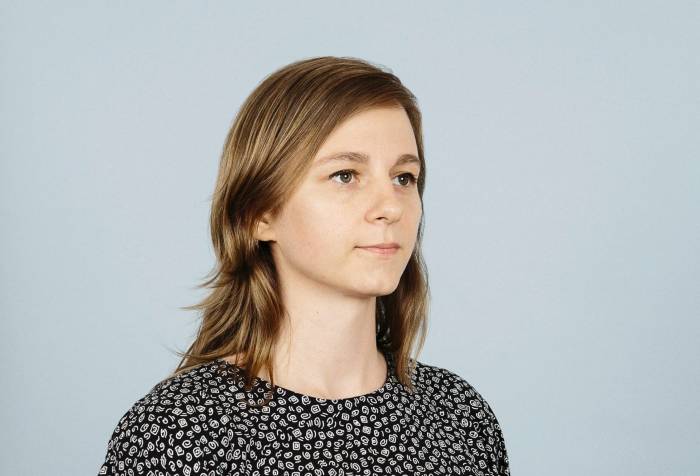 In this portrait, I’m facing askew from the camera and smiling slightly. I have medium-length wavy blonde hair, and I’m white. I’m 30, but people often tell me I look younger. I’m wearing a shirt with a pattern of white squiggles on black paired with bright blue dress pants.