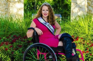 A young woman in a sleeveless hot pink dress and black leather boots. She is sitting in a wheelchair and wears a crown. She also has a sash that says "Ms. Wheelchair Florida."