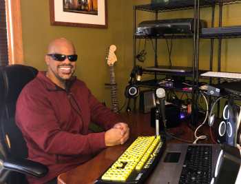 Thomas Reid an African American man with a clean shaven head and goatee in shades smiles at the camera while seated at a desk. The desk holds a laptop and other equipment including an audio mixer and microphone. A framed picture of the original World trade Center hangs on the wall above a black Fender electric guitar.