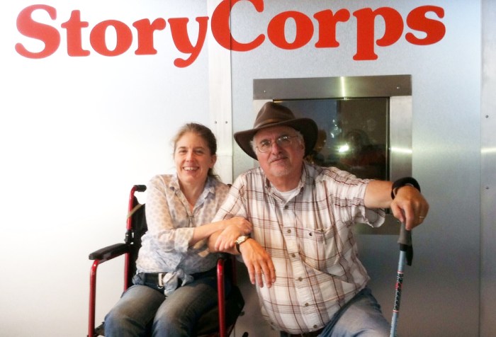 Picture outside a metal recording booth with a sign in the background that reads 'StoryCorps' in the front is a middle-aged woman in a wheelchair. She is clasping the arm of an older man who is next to her, bending his knees halfway down. He is wearing a hat and holding on to a walking stick.