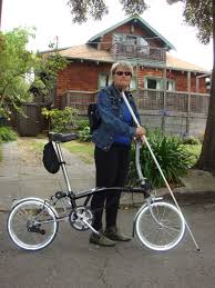 White woman wearing dark sunglasses holding a white cane. She is standing outside of a house and holding a bike.