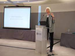A woman with white hair and a gray blazer with black pants standing at a lectern. She is holding a white cane and screen is to the left of her showing a slide.