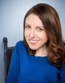 Woman with brunette hair in blue top, sitting in a wheelchair.