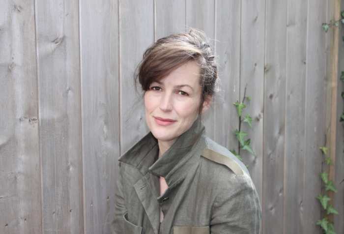 Sara Hendren, a white middle aged woman with brown hair and brown eyes, sits in front of a weathered wood fence, wearing an olive linen jacket.
