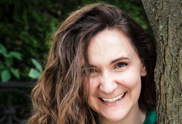 A young white woman with long curly brown hair and brown eyes smiles at the camera. She stands beside a brown tree trunk and is wearing an emerald green tunic top and a white cardigan sweater.