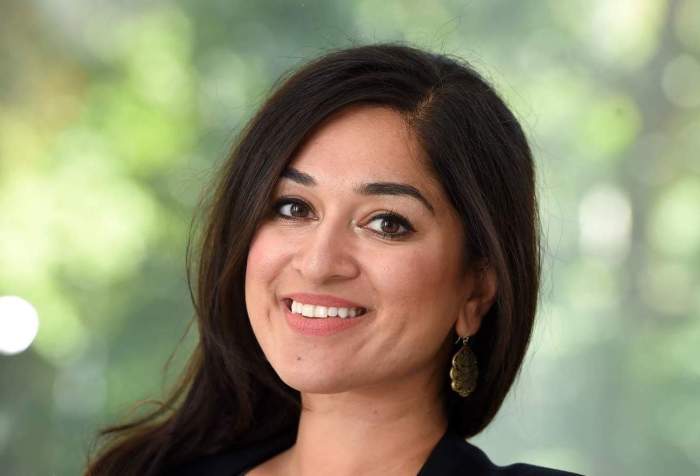 Jaipreet Virdi, a South Asian woman with dark hair, smiles at the camera. Her head is slightly turned. She is wearing a black blazer and dark top. There are some trees in the blurred background.
