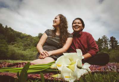 A photo of Elena and Layel smiling and looking off beyond the camera. They are both sitting on a cloth with calla lilies with an evergreen forest behind them.