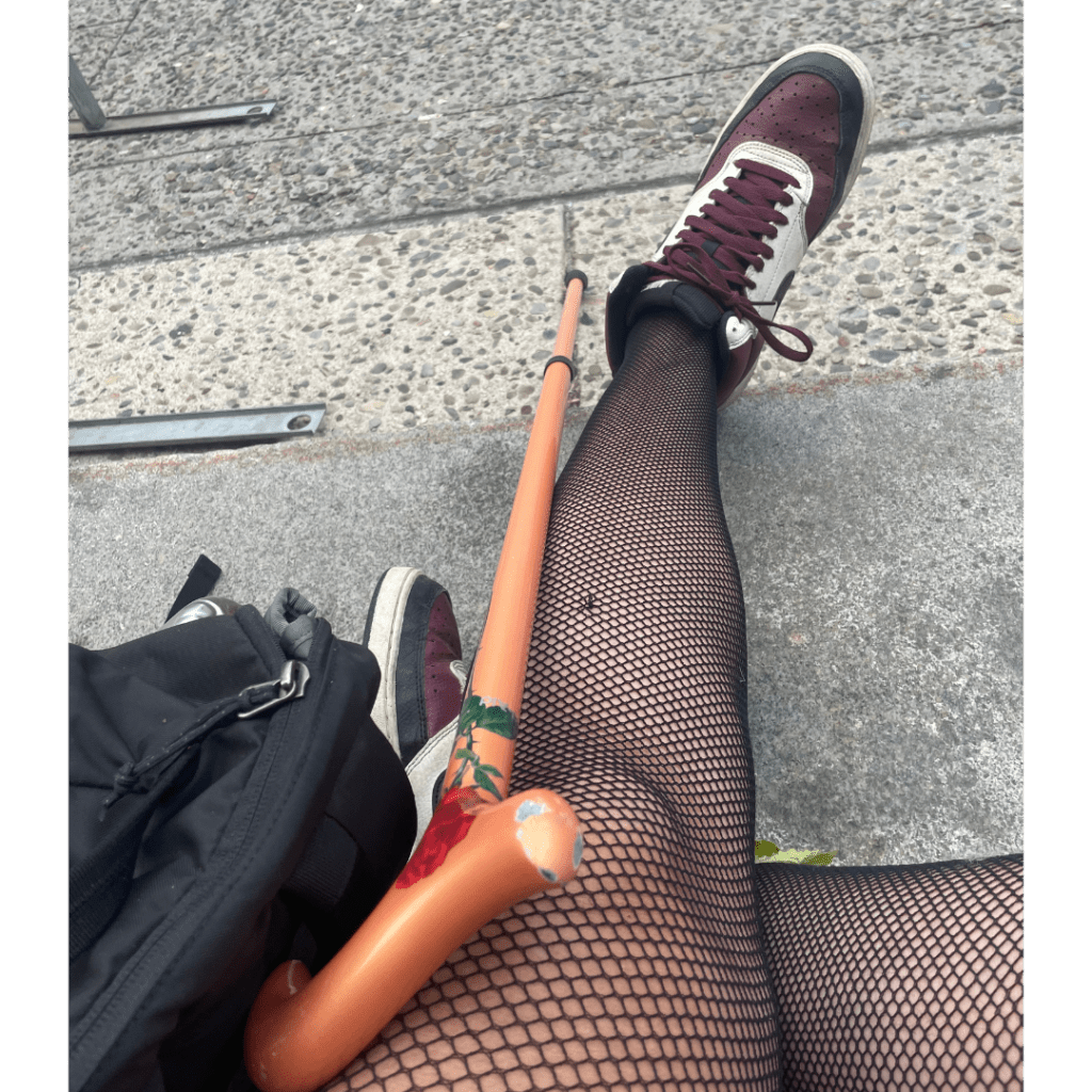 A photo looking downward at Leah's crossed legs as they sit down on grey stone steps (post a march, watching speakers, though you can't see that). Their legs are fishnets, a white, brown and black high top sneaker and their rose gold cane with a red rose are visible and a black backpack is to one side.