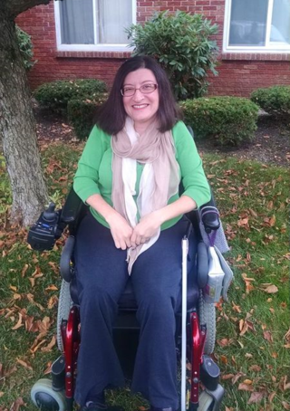 Middle-aged white woman with long brown hair and glasses sitting in a wheelchair outside of her home. She is wearing jeans and a green sweater.