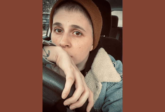 Graphic with a brown background with a photo of A Andrews, a white non-binary person with brown eyes and short hair. They are sitting in the driver’s seat of a car with dark seats, wearing a tan beanie, and lined denim jacket.