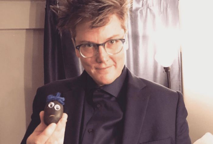 Image is a photo of Hannah Gadsby in a dark suit, holding some of her handmade pet rocks.